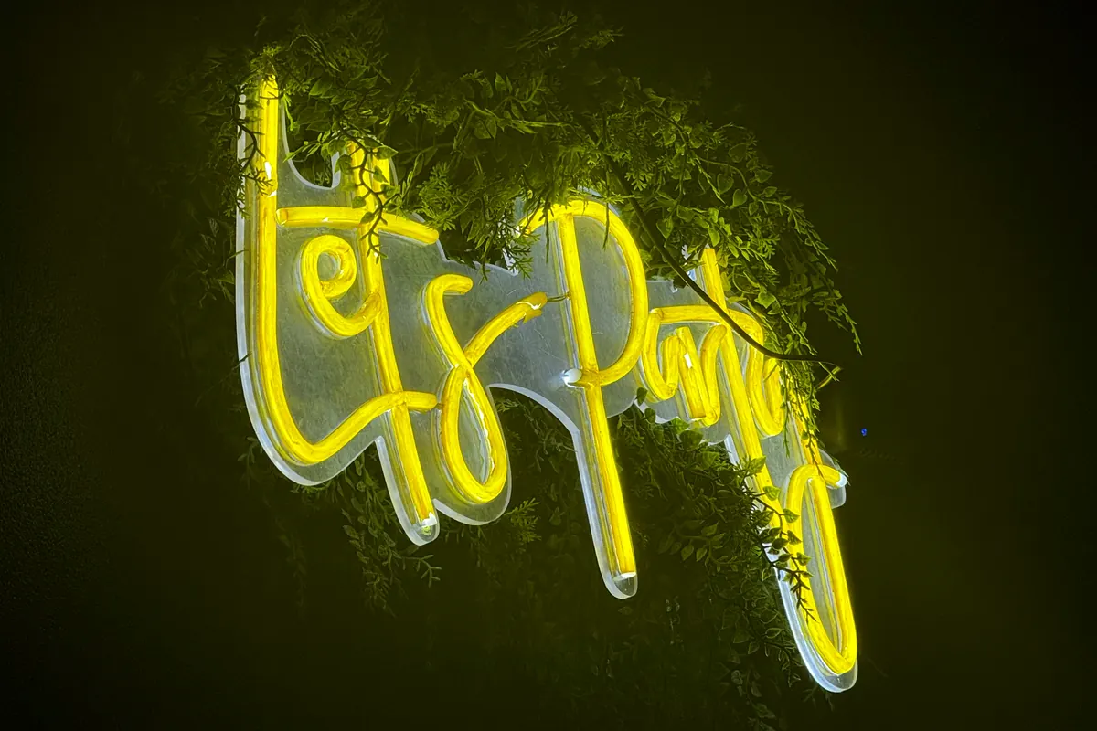 Rótulo de neón amarillo que dice «Let's Party» sobre la pared de plantas.